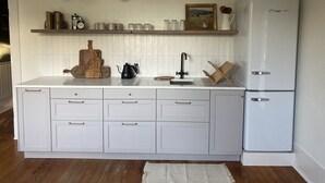 Une cuisine avec des meubles blancs, un réfrigérateur blanc, une planche à découper en bois, une cafetière et un luminaire suspendu.
