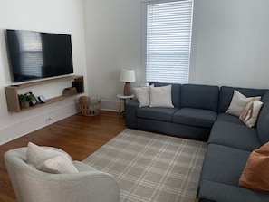 A living room with a flat-screen TV, a grey sofa, a small round table with a lamp, and a wicker basket. - Historic • Hot tub • A&M Ring Day (Bryan)