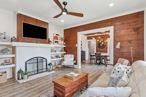 A living room with a fireplace, a television mounted above it, and a dining area visible through an open doorway.