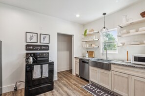 A modern kitchen with a built-in oven, dishwasher, and a stainless steel sink. There are framed botanical prints on the wall and a kitchen sign that reads 'cook, sing, dance.'