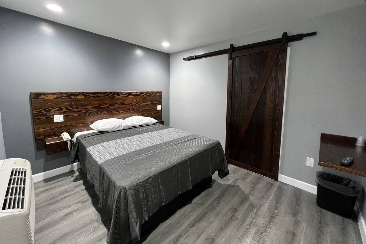 Un dormitorio con cama, mesita de noche, calefactor, puerta corrediza tipo granero y un cubo de basura.