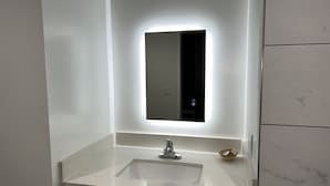 Un baño con lavamanos blanco, un espejo con iluminación trasera y una pequeña cesta de mimbre en la encimera.