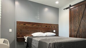 Un dormitorio con cabecera de madera, cama con colcha estampada, una silla blanca y un armario de madera.
