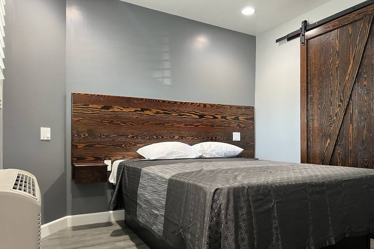 Un dormitorio con cabecera de madera, cama con colcha estampada, una silla blanca y un armario de madera.