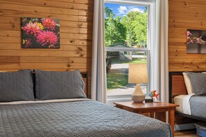 A bedroom with a bed, a nightstand, a lamp, and a window with a view of greenery. - Private Hot Tub!  Kayaks. Serene Lakefront. Pool. (Forsyth)