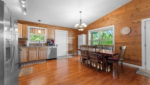 A kitchen with wooden cabinets, a dining table with chairs, a chandelier, and a clock on the wall.