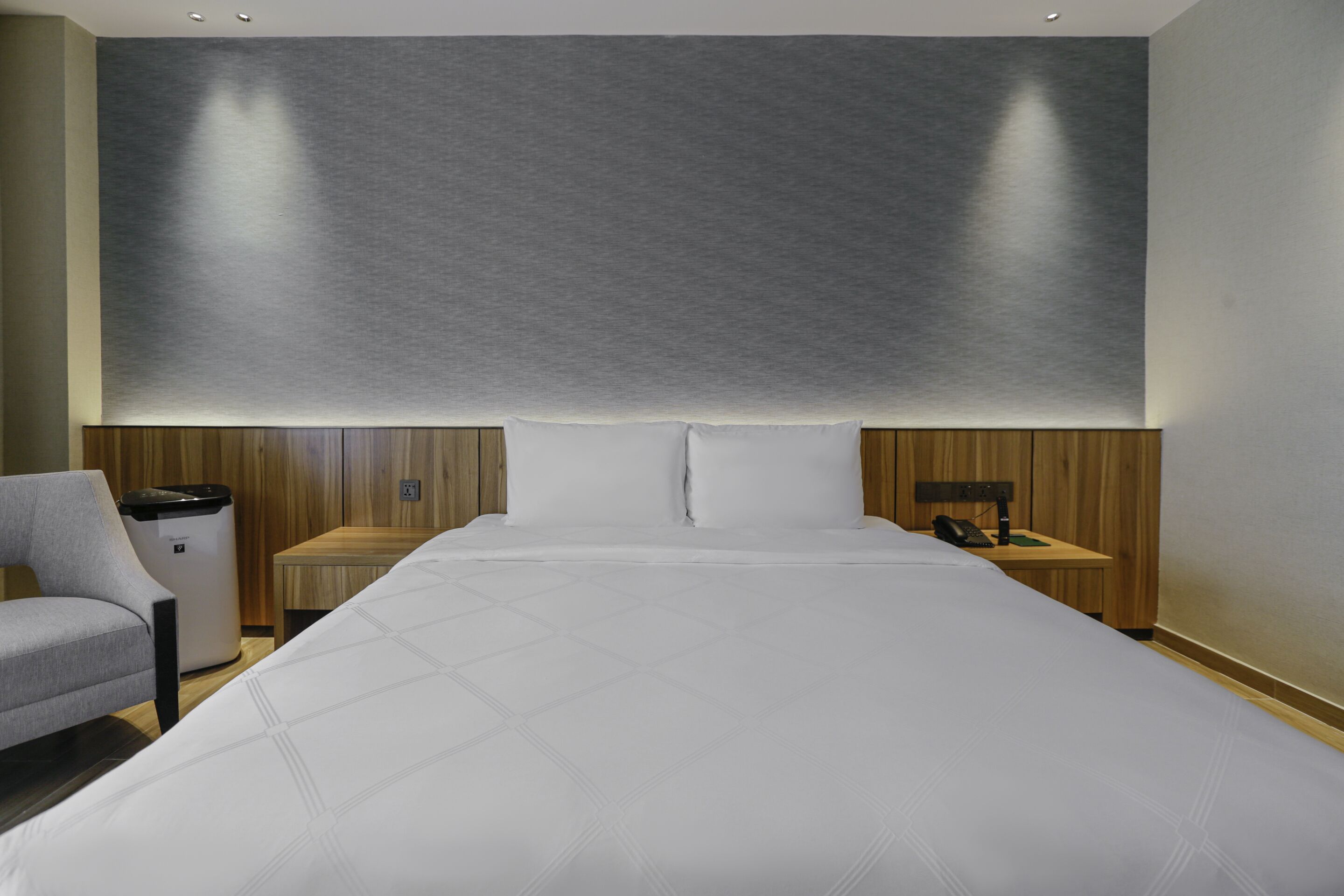 A modern hotel room with a large bed, a wooden nightstand, a chair, and a wall-mounted light fixture.