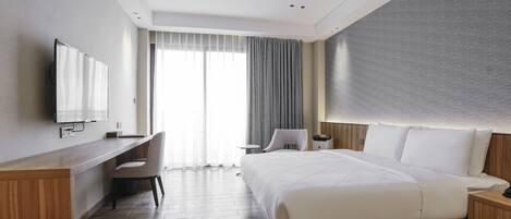 Una habitación de hotel moderna con un escritorio de madera, una cama con ropa blanca, un televisor montado en la pared y un amplio ventanal con cortinas.