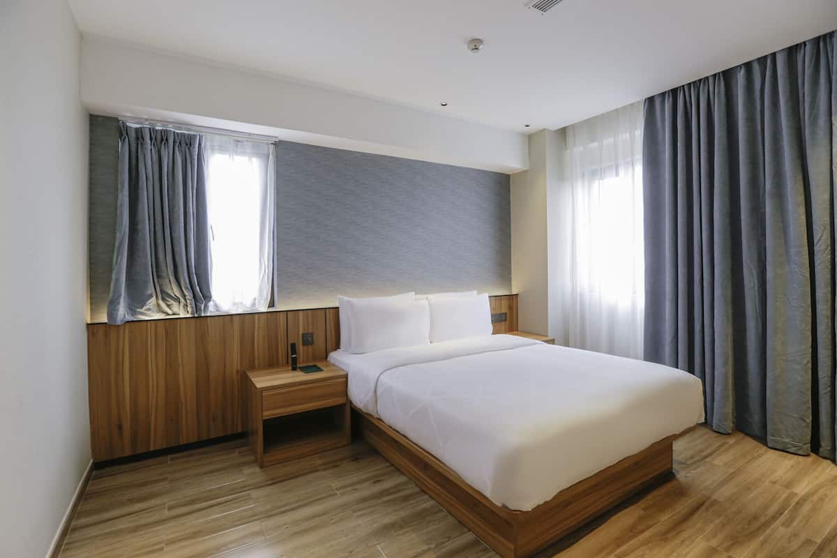 A modern hotel room with a wooden headboard, a large bed, a nightstand, and a window with curtains.