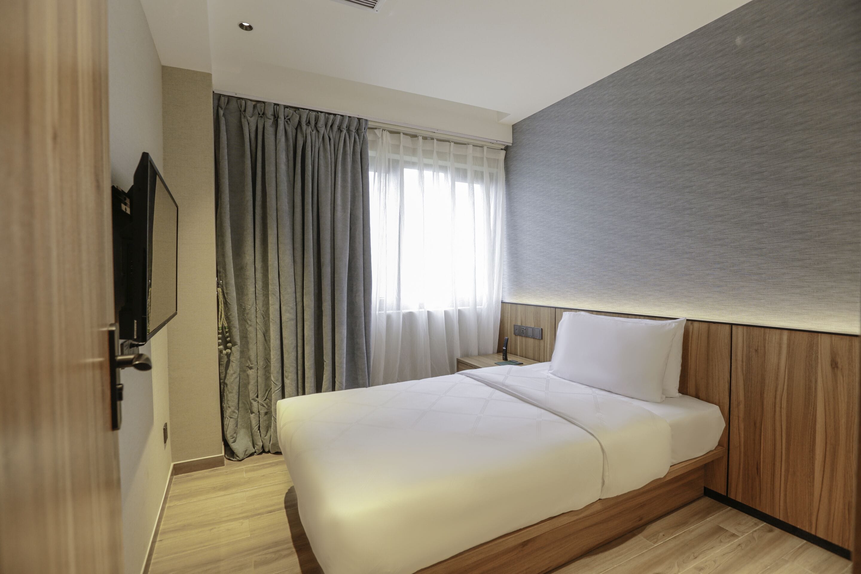 Habitació d'hotel amb llit, televisió, finestra amb cortines i sòl de fusta.