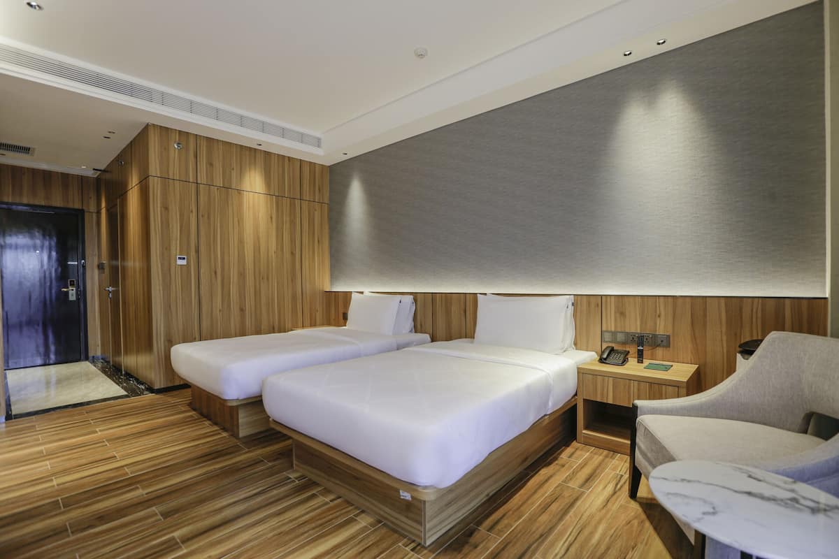 A modern hotel room with two single beds, a wooden floor, a large mirror, a small desk with a telephone, and a grey armchair.