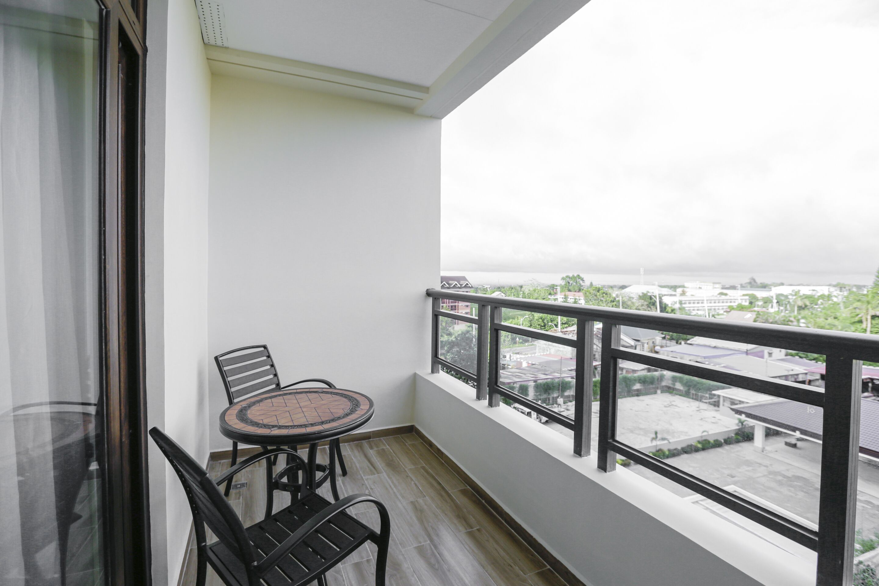 A balcony with a table and chairs, a glass door, and a view of buildings and greenery.