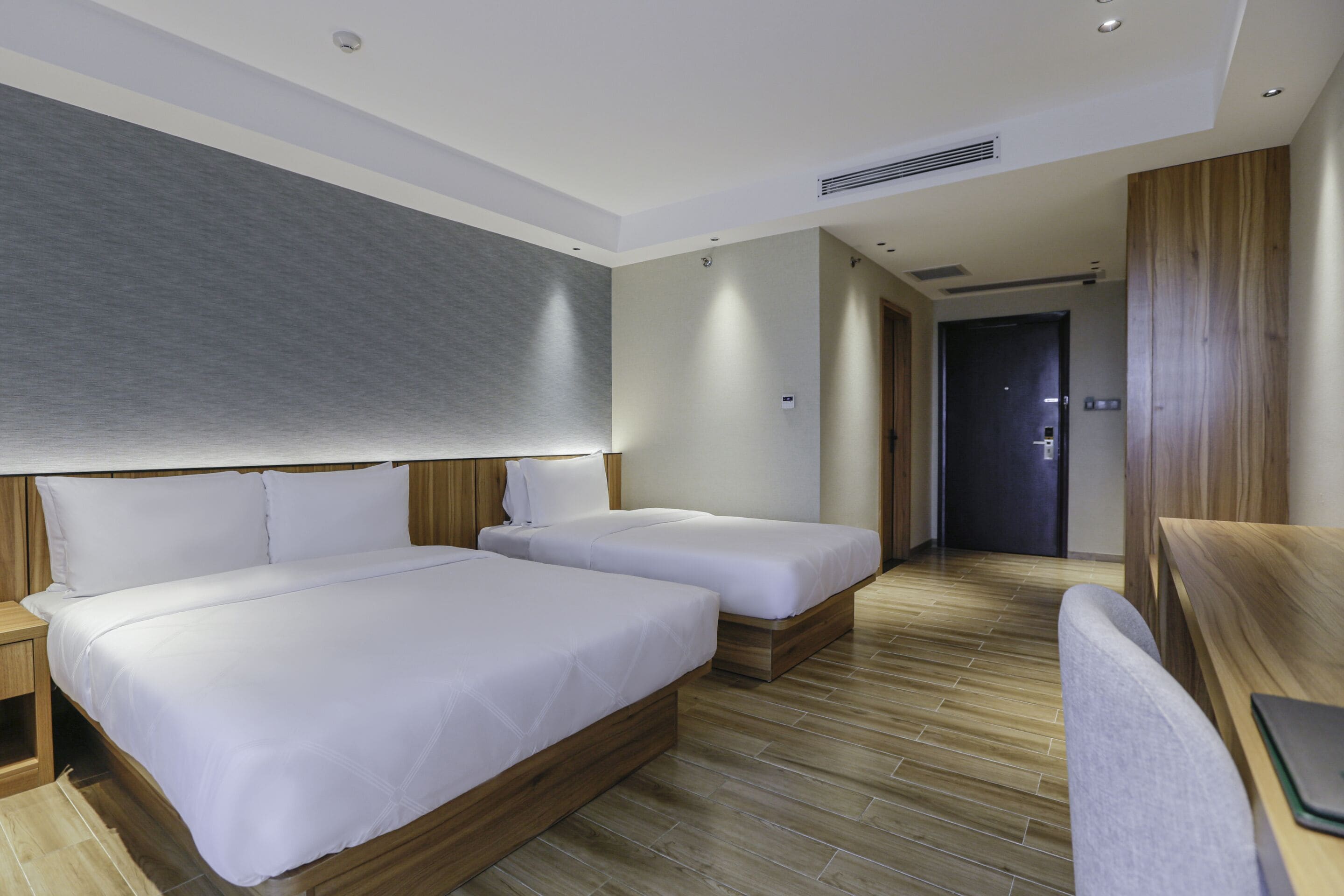 Habitación de hotel con dos camas individuales, cabecero de madera, escritorio con silla y puerta que da a otra habitación.