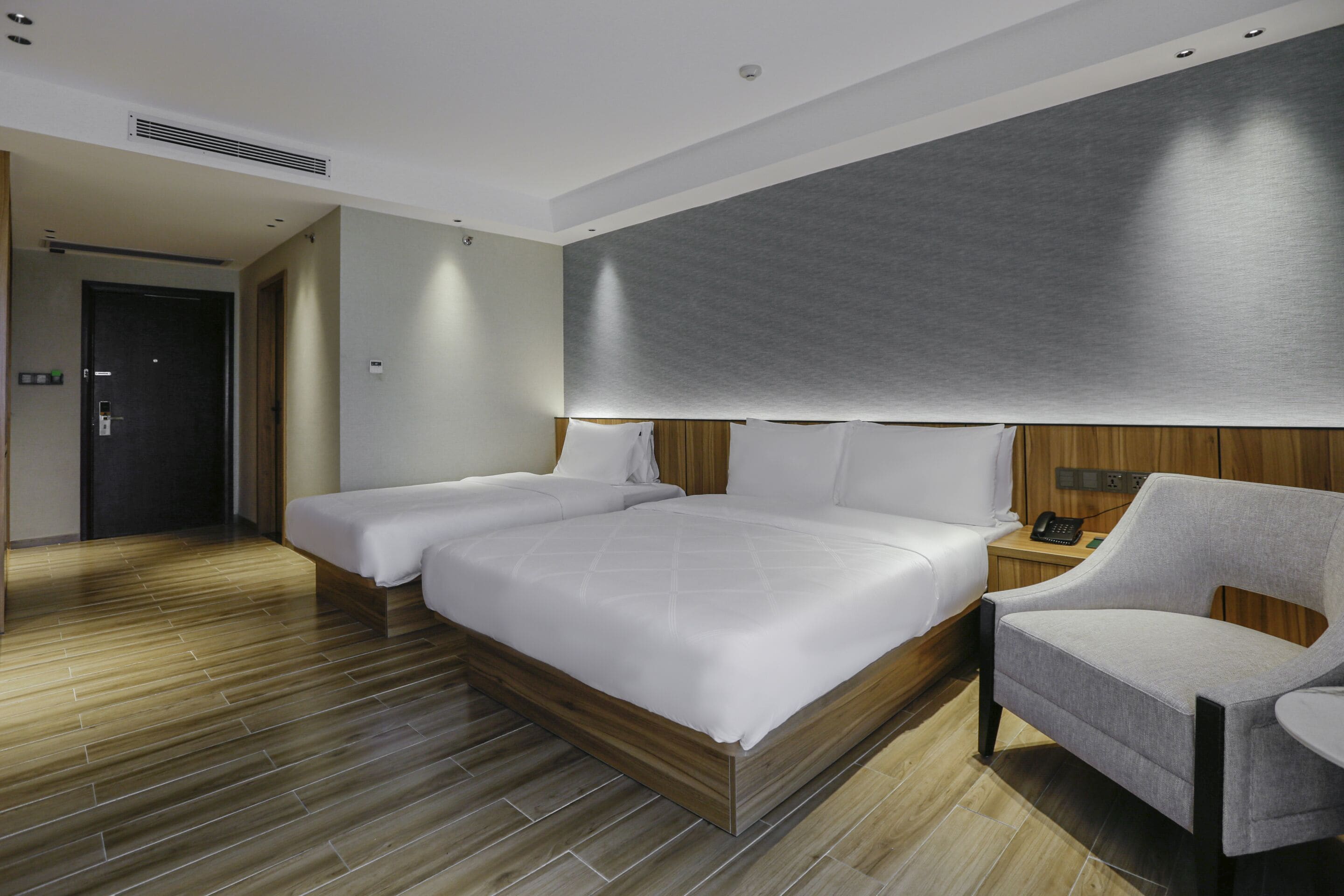 A modern hotel room with a large bed, a bedside table with a lamp, and a wooden floor.