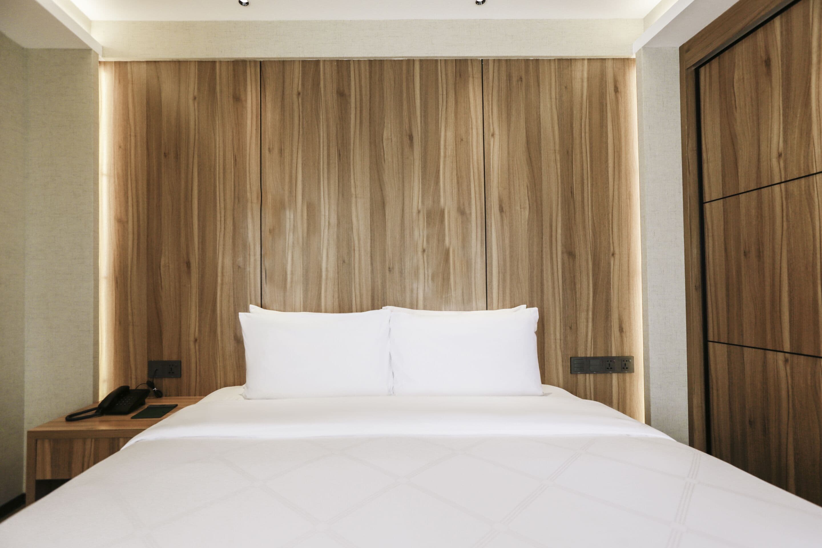 A hotel room with a wooden headboard, a bed with white bedding, a bedside table with a phone, and a nightstand.
