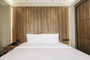 Habitación de hotel con cabecera de madera, cama con ropa de cama blanca, mesita de noche con teléfono y velador.