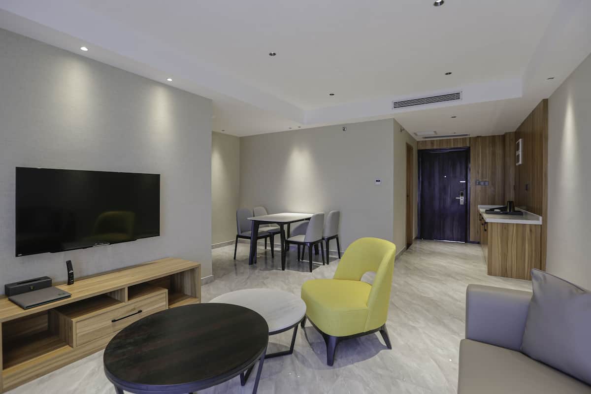 A modern living room with a flat-screen TV, a round coffee table, a yellow armchair, a grey sofa, and a dining area with a table and chairs.