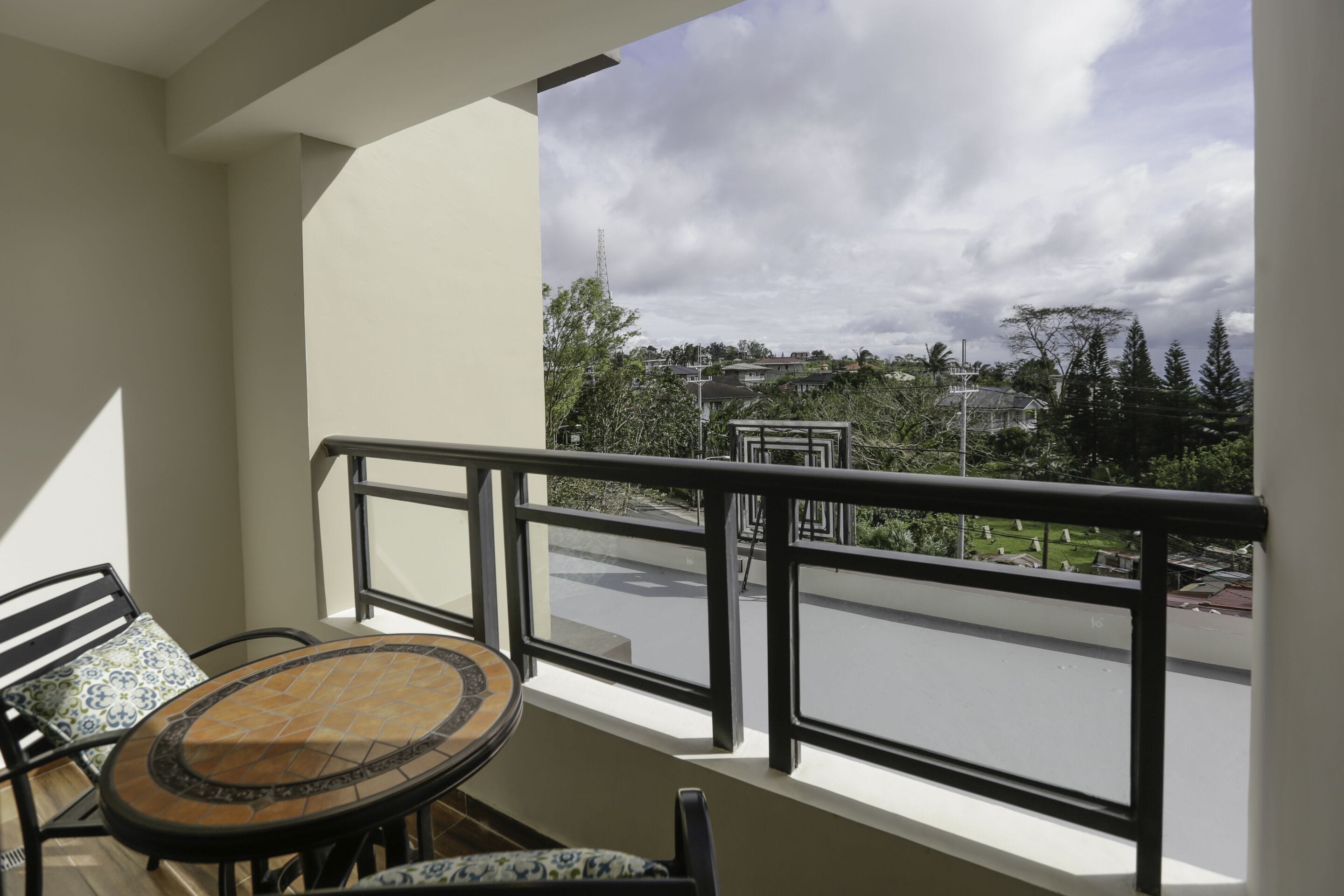 A balcony with a round table and chairs, a view of a residential area, and a cloudy sky.