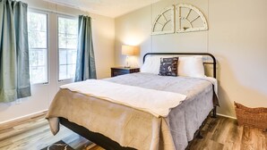 Quarto com cama, mesa de cabeceira, abajur e janela com cortinas.