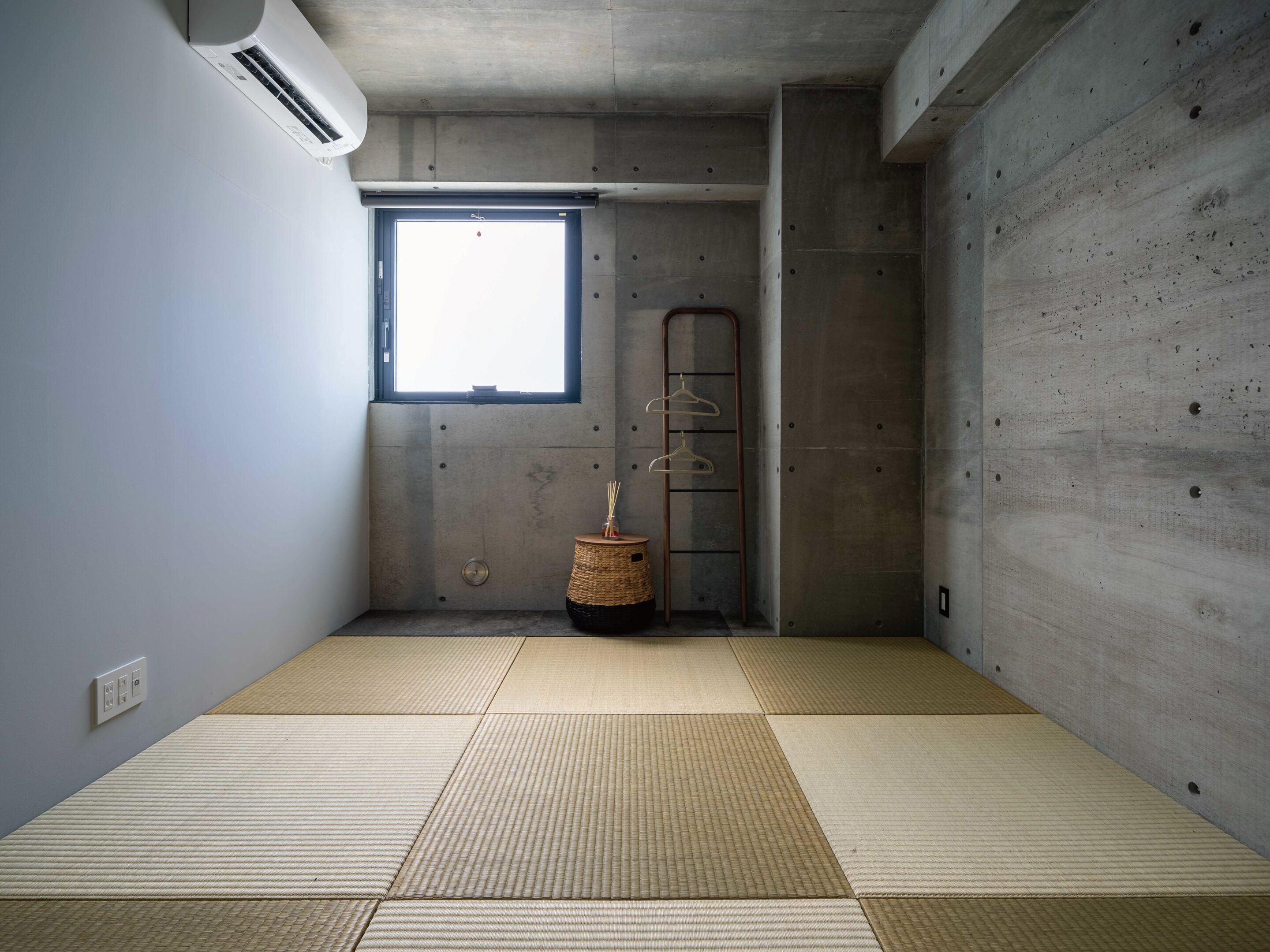 A room with tatami mats, a concrete wall, a window, and a hanging rack.