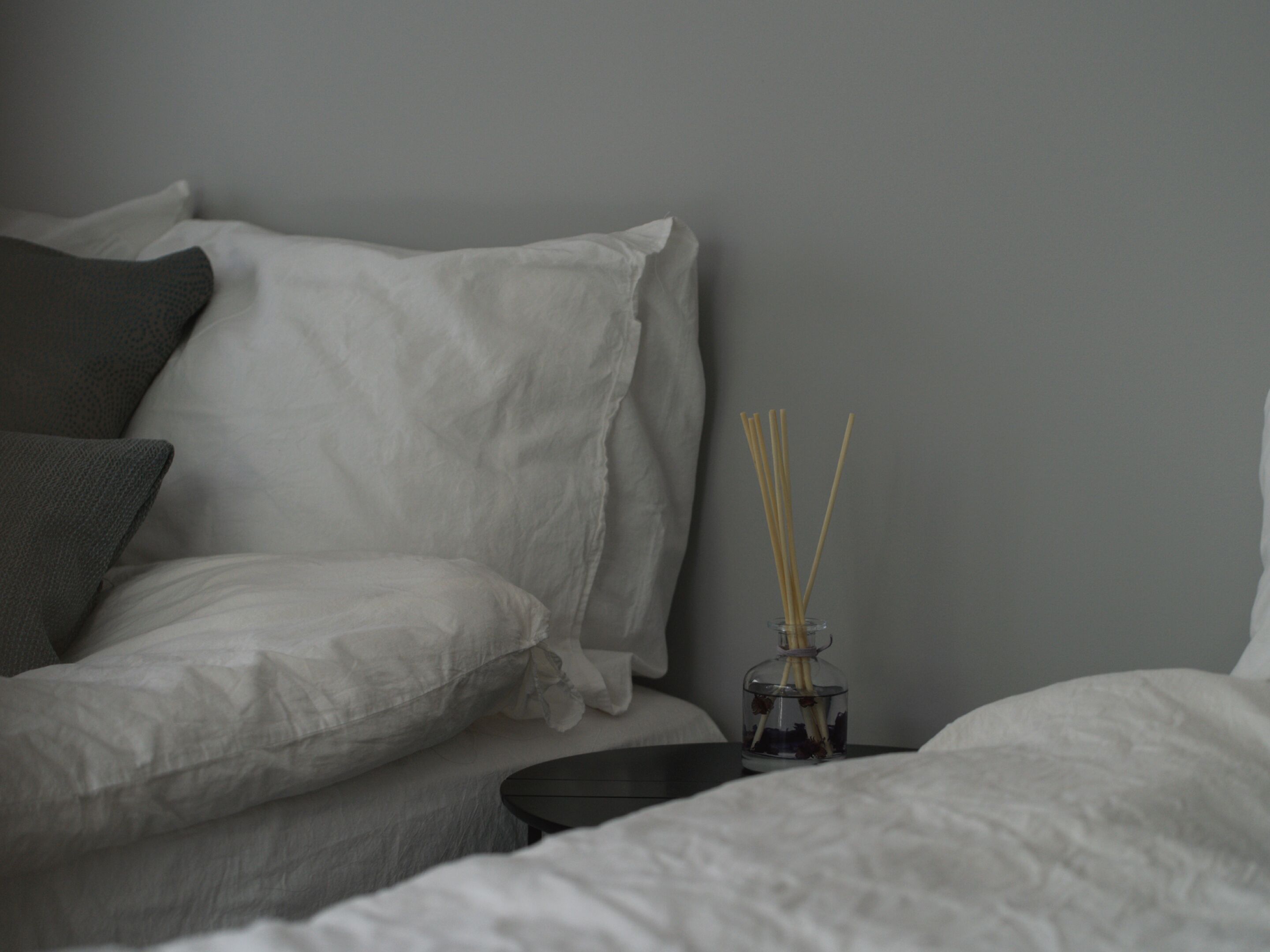 Een net opgemaakt bed met wit en grijs beddengoed, een klein zwart nachtkastje en een glazen vaas met rietstelen.