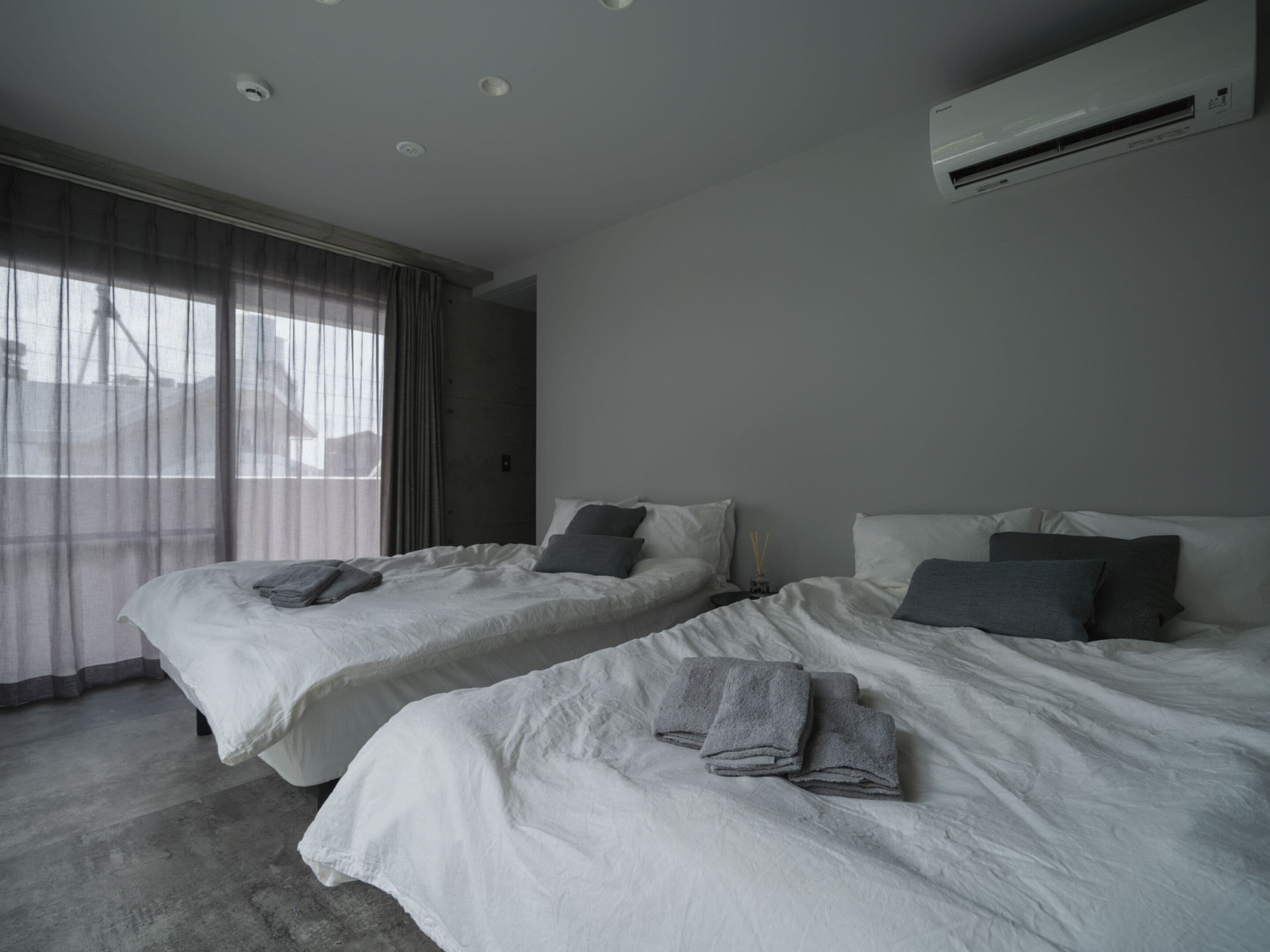 A hotel room with two beds, a large window with sheer curtains, and an air conditioning unit on the wall.