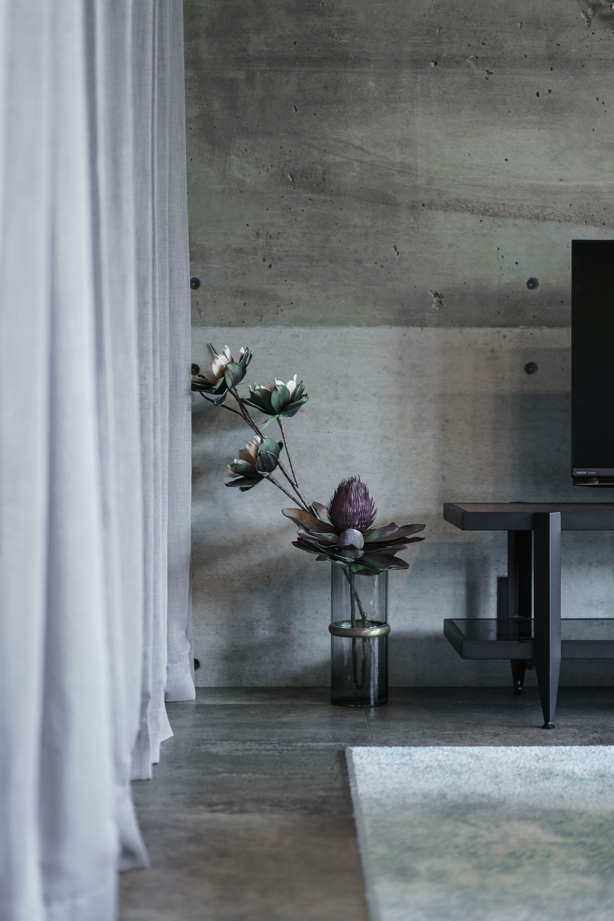 A modern room with a concrete wall, a small glass vase with flowers, a dark wooden table, and a white curtain.