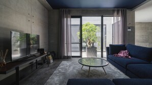 Un salon moderne doté d’un téléviseur à écran plat, d’une table basse en verre, d’un canapé bleu et d’une grande fenêtre donnant sur un paysage verdoyant.