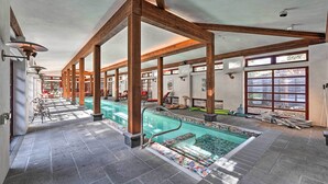 En indendørs swimmingpool med træbjælker, flisegulv og udsigt til det fri gennem store vinduer.