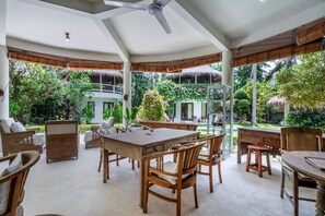 Un amplio espacio de comedor al aire libre con mobiliario de madera, un muro de cristal que ofrece vistas a exuberante vegetación y un edificio.