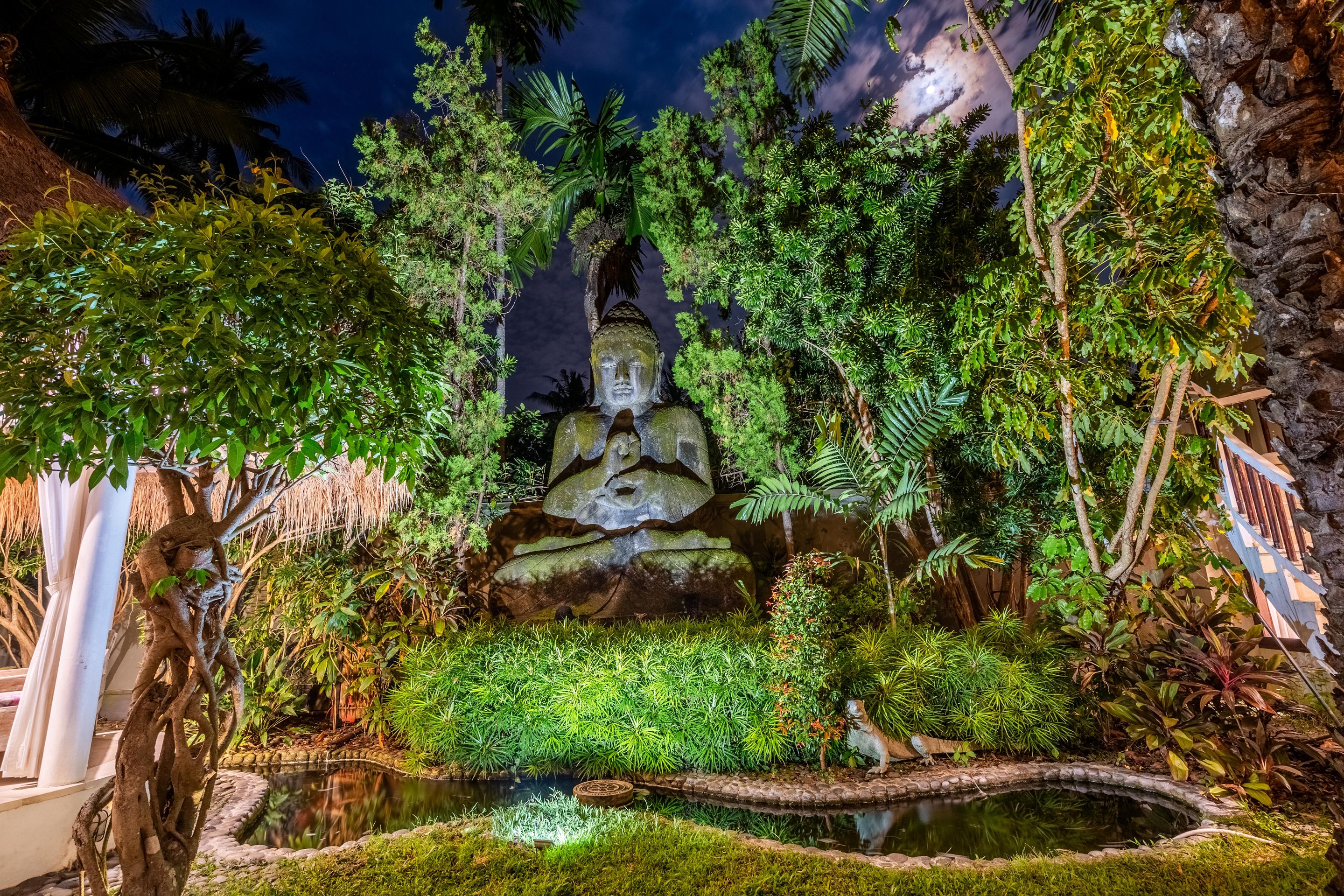 Um jardim tranquilo com uma estátua de Buda, vegetação exuberante e um pequeno lago.