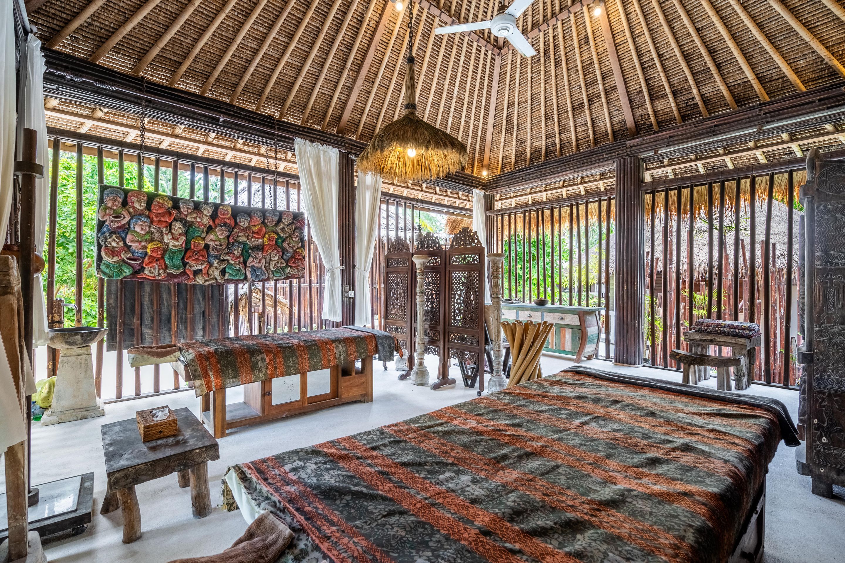 Quarto tradicional com telhado de palha, cama com colcha estampada, mesa de madeira e parede com arte colorida.