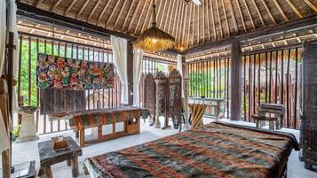 Quarto tradicional com telhado de palha, cama com colcha estampada, mesa de madeira e parede com arte colorida.