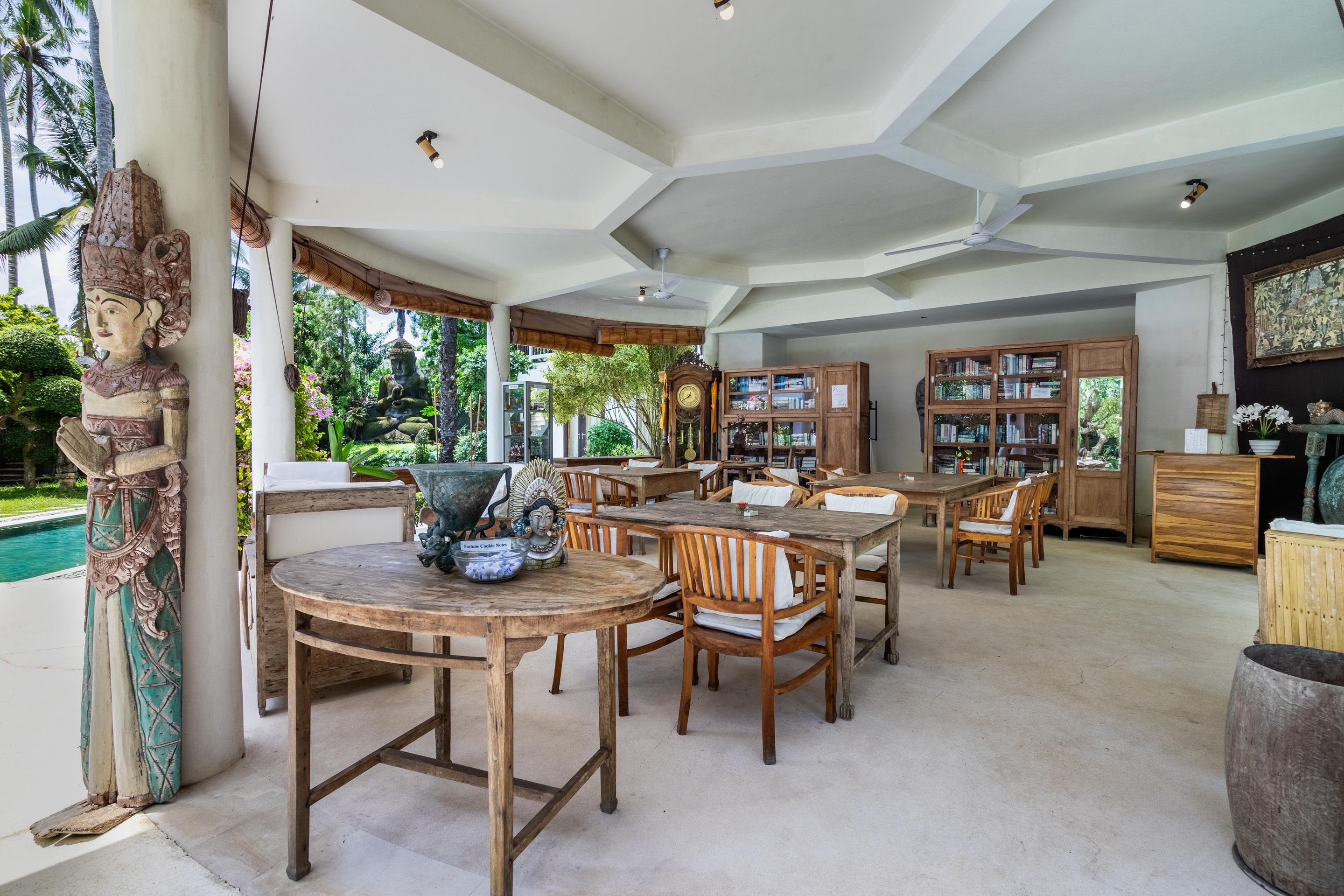 A spacious dining area with wooden tables and chairs, a large Buddha statue, and a view of a lush garden.
