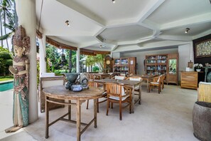 A spacious dining area with wooden tables and chairs, a large Buddha statue, and a view of a lush garden. - Double Room with Pool/Garden View | The Village of Angels Resort (Kabupaten Karangasem)