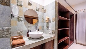 Un baño con pared de piedra, un espejo redondo, un lavabo blanco y estantes de madera.