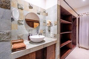 Un baño con pared de piedra, un espejo redondo, un lavabo blanco y estantes de madera.