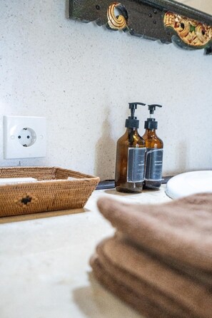 Une salle de bain avec un lavabo blanc, deux bouteilles marron étiquetées « gel douche » et « shampoing longue durée », une prise de courant blanche et un panier en osier.