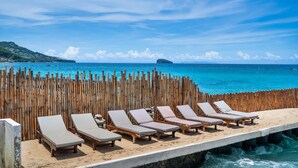 Vista de la playa con sillones de madera alineados a lo largo de un borde de hormigón, con vista a un mar azul cristalino y una isla distante.