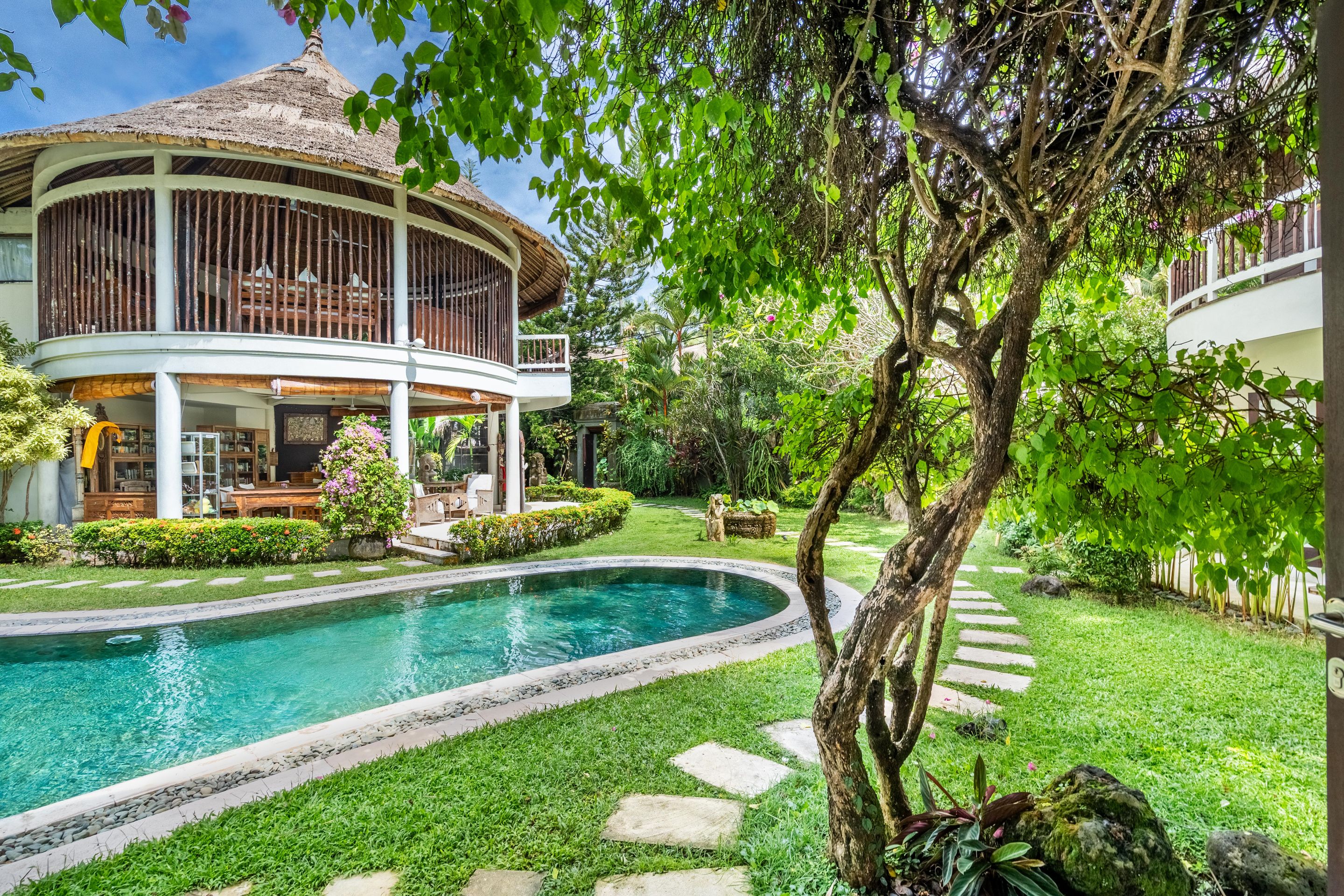 A pool surrounded by a well-maintained garden, a building with a thatched roof, and a tree with hanging branches.