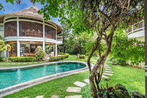 A pool surrounded by a well-maintained garden, a building with a thatched roof, and a tree with hanging branches.