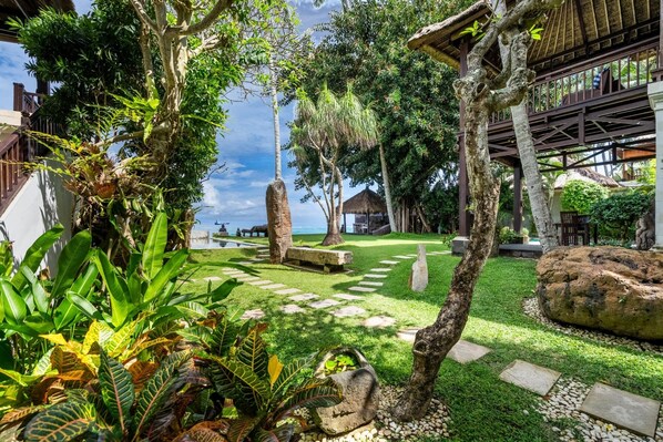 Un jardin tropical avec un sentier en pierre, des palmiers et une structure surmontée d’un toit de chaume.