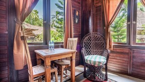 A wooden room with a table and chairs, a window with curtains, and a view of greenery outside. - Double Room with Alley View | The Village of Angels Resort | Candidasa (Kabupaten Karangasem)