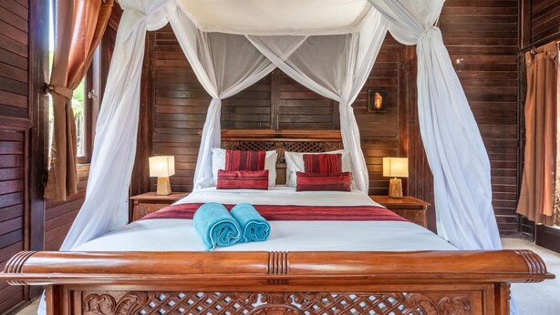 A wooden four-poster bed with a canopy, red and white bedding, and two blue towels. - Double Room with Alley View | The Village of Angels Resort | Candidasa (Kabupaten Karangasem)