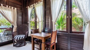 A wooden room with a table and chairs, large windows with curtains, and a view of greenery outside. - Deluxe Double Room with Balcony and Ocean View | The Village of Angels Resort (Kabupaten Karangasem)