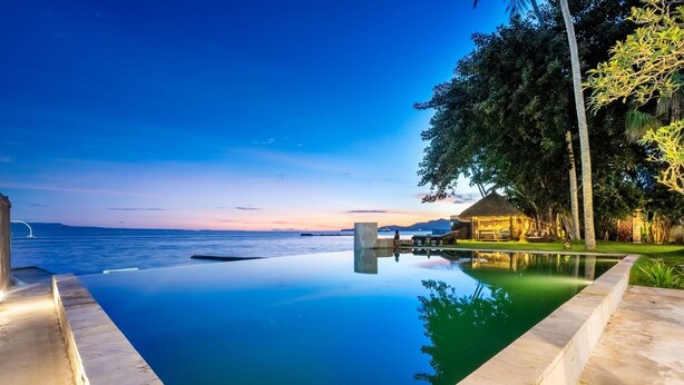 A infinity pool with a view of the sea and a thatched-roof structure.