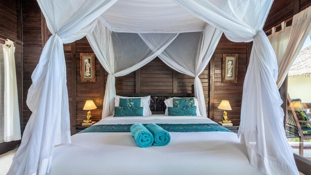 A four-poster bed with white drapes, teal pillows, and a rolled towel on the bed. - Deluxe Double Room with Balcony and Ocean View | The Village of Angels Resort (Kabupaten Karangasem)
