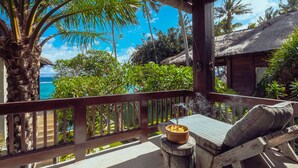 A wooden deck with a table and chairs, overlooking a beach and palm trees. - Deluxe Double Room with Balcony and Ocean View | The Village of Angels Resort (Kabupaten Karangasem)
