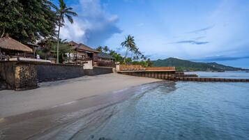 A beachfront property with a sandy beach, a wooden pier, and thatched-roof buildings.