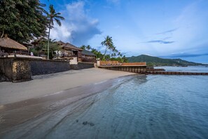 A beachfront property with a sandy beach, a wooden pier, and thatched-roof buildings. - Beachfront Group Property – 9 Separate Rooms, 3 Levels | The Village of Angels (Kabupaten Karangasem)
